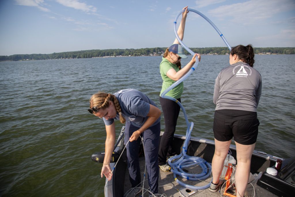Hyrcik and Yerofeev collect water to wash any contaminants out of the containers that will hold water samples from Chautauqua Lake, so as to not obstruct the data, while Beth Fisher collects samples on the other side of the boat.